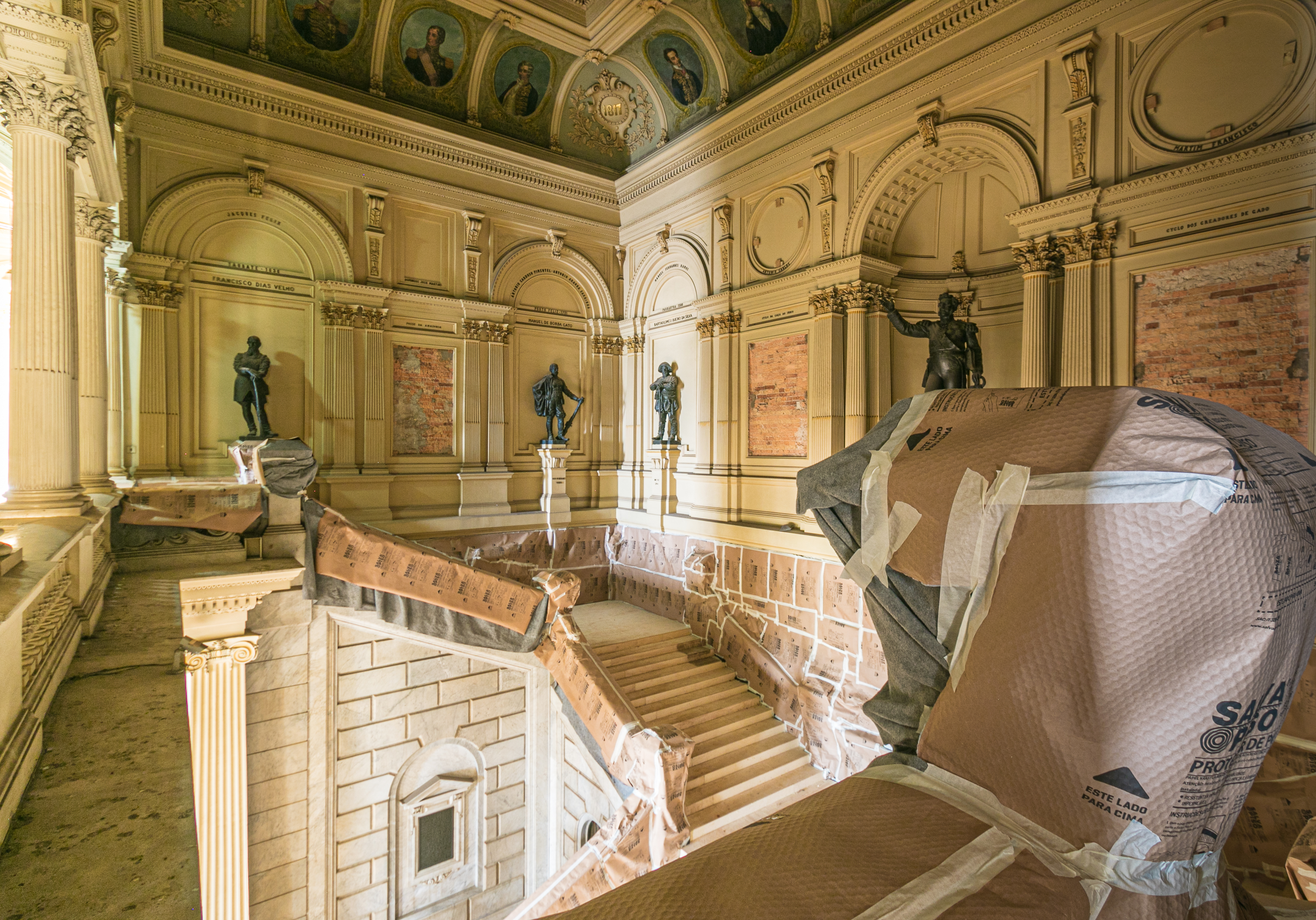 Audiodescrição: Foto de parte da escadaria e do segundo andar do Museu Paulista. À frente está a escadaria da lateral esquerda. Algumas paredes, os corrimões e o corredor estão cobertos com papéis de proteção. Há algumas estátuas expostas.